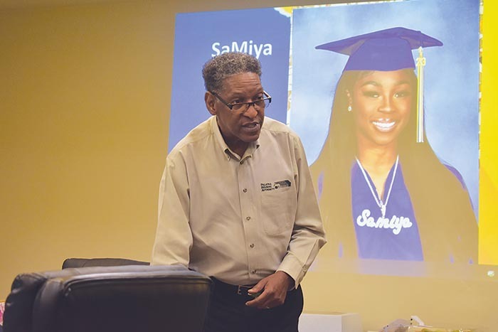 Resident Services Director reaching under podium to retrieve an item while in front of a projector displaying a recent female graduate 